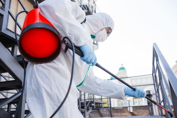 A person in a white protective suit applies pest control spray to a red object, ensuring effective treatment against pests.