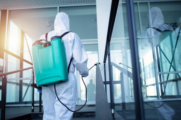 A person in a white protective suit using a sprayer for pest control in an outdoor setting.